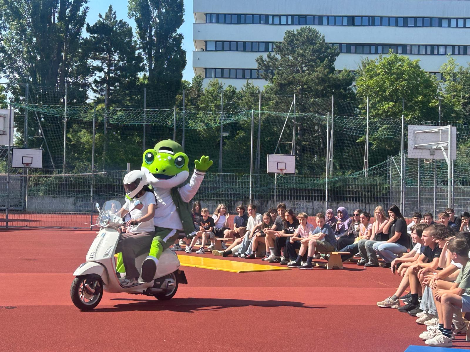 High quality mascot costume of a green frog riding on a scooter with a driver in front of a seated crowd of students at an outdoor sports court event.