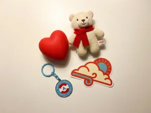 Branded promotional items including a plush bear, stress ball, keychain, and custom sticker arranged in a flat-lay on a white surface.
