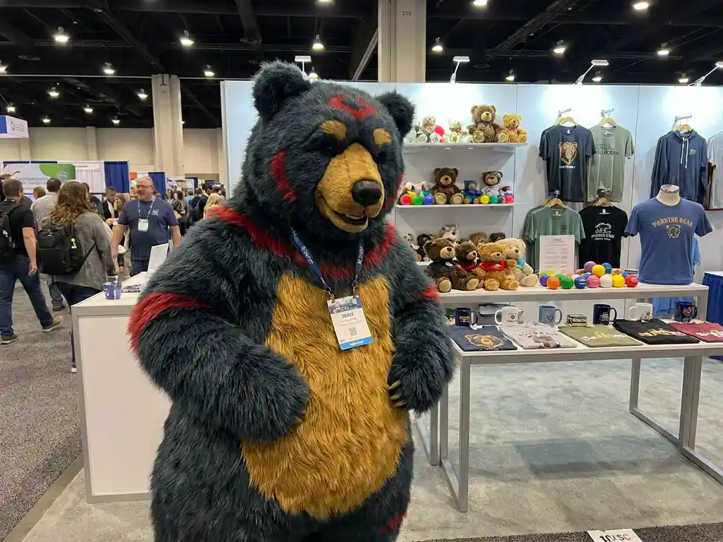 Custom bear mascot costume at a trade show booth displaying plush toys, stress balls, and branded merchandise under bright expo lighting.